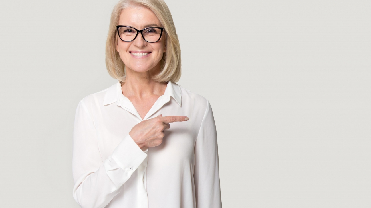 Happy old woman in glasses showing to copyspace blank space. Smiling elderly female looking at camera holding finger point to left to empty place for text. Studio portrait isolated on gray background