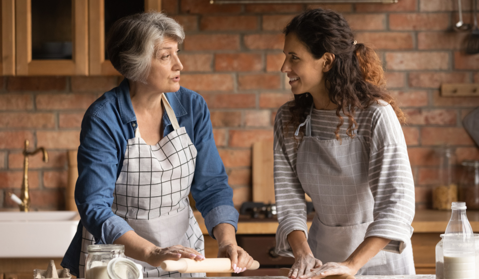Mature grey haired woman with grownup daughter chatting, cooking homemade pastry, rolling dough, standing in kitchen at home, family spending leisure time together, preparing pie or bread together