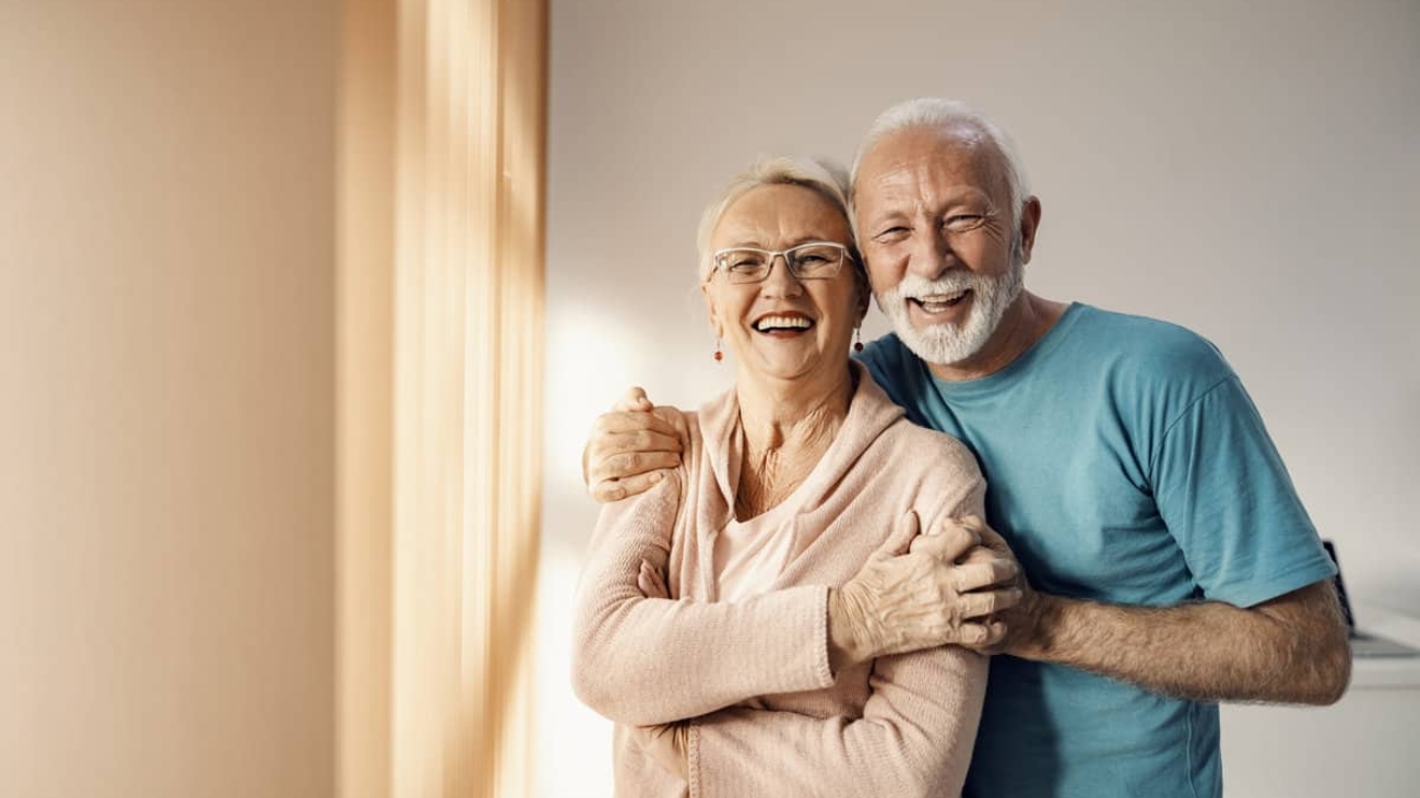 Senior couple hugging in a nursing home. A happy senior couple standing next to a window in a nursing home, hugging and smiling. They have all care they need.
