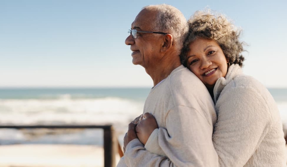 Romantic senior woman smiling at the camera while embracing her husband by the ocean. Affectionate elderly couple enjoying spending some quality time together after retirement.