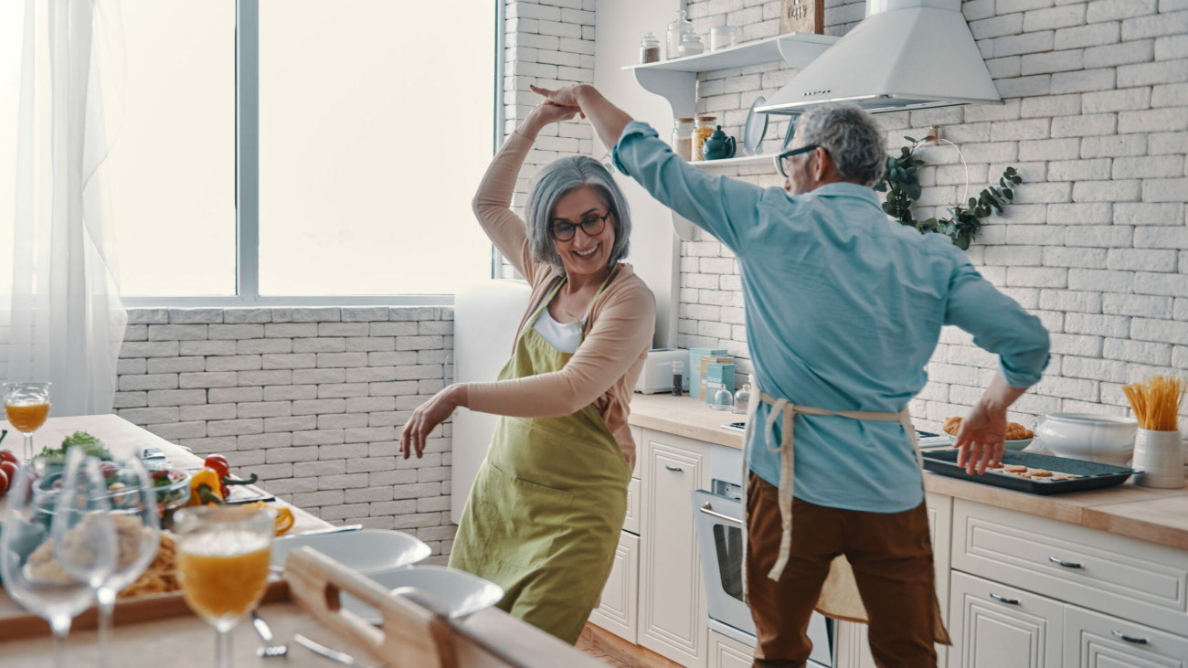 Beautiful playful senior couple in aprons dancing and smiling while preparing healthy dinner at home